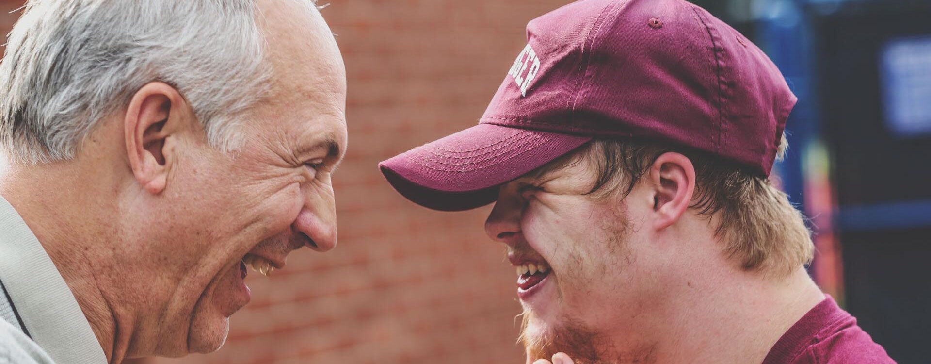 Dwóch mężczyzn patrzy na siebie. Starszy uśmiecha się do młodszego. Młodszy ma widoczne objawy zespołu Downa.